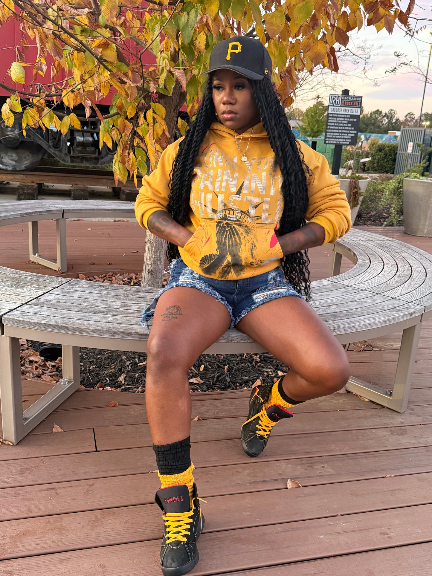 Lady wearing my yellow hoodie with rhinestones on front Statue of Liberty and blue shorts sitting on a wooden bench with autumn leaves.