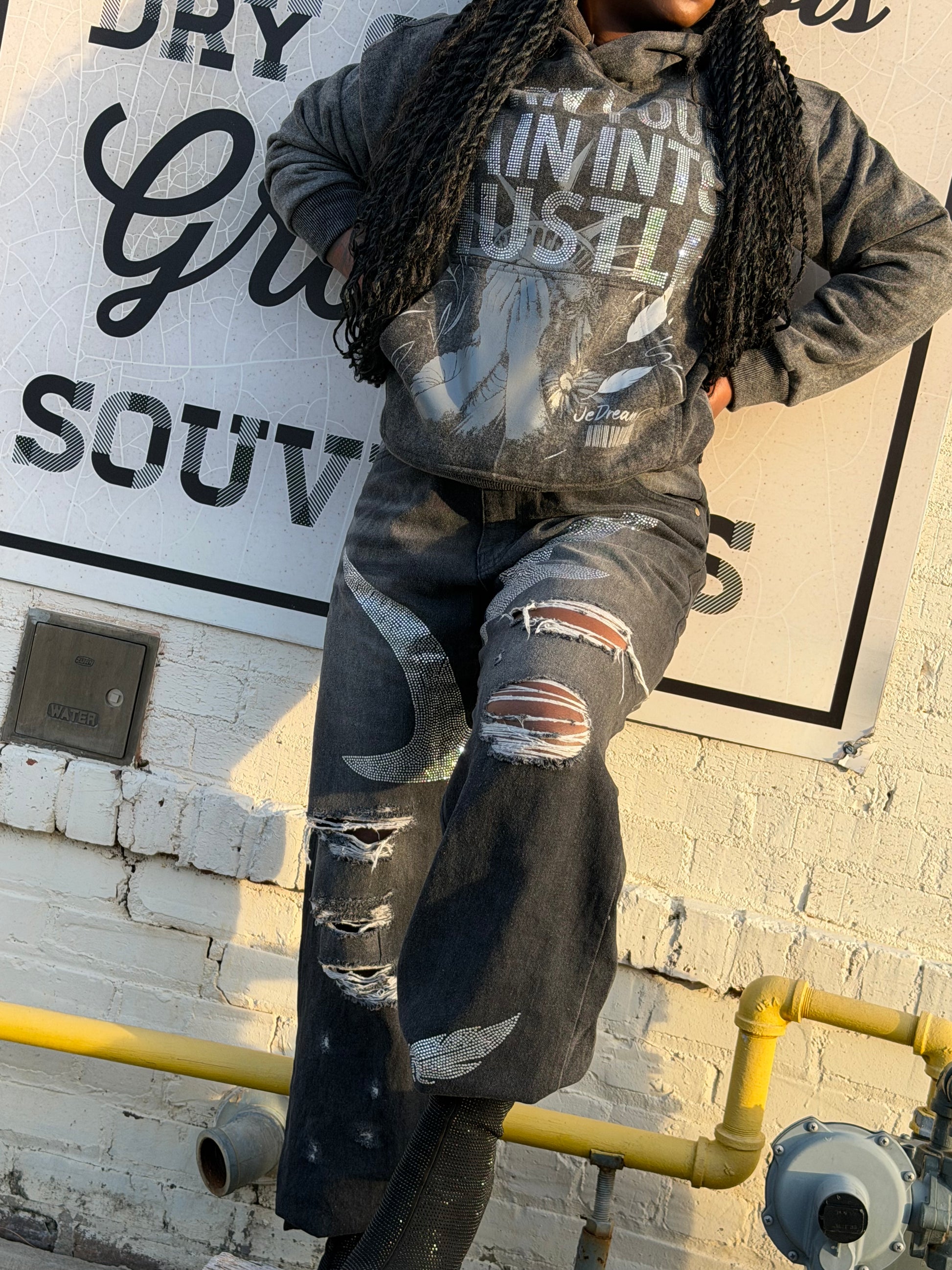 Lady wearing a distressed black jeans with rhinestones and black acid hoodies a white wall with black text.