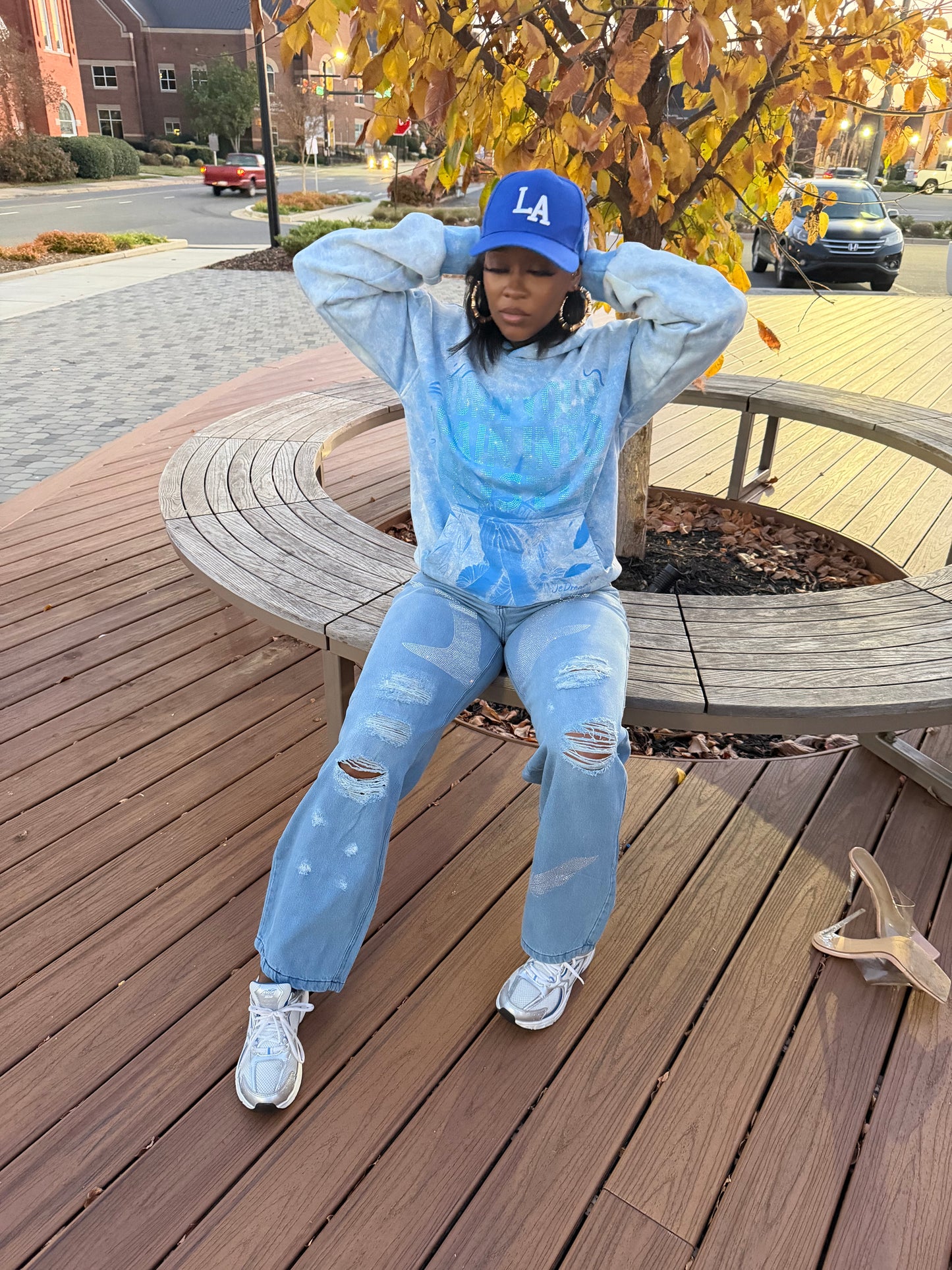 Lady wearing my light blue acid wash hoodie sit on a wooden bench showing off the hoodie and wearing our je dream co rhinestones jeans 