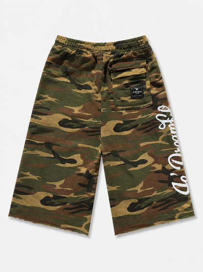 A pair of camouflage shorts in green, brown, and tan, featuring a white script logo printed vertically on the left leg and a black brand patch on the back pocket.