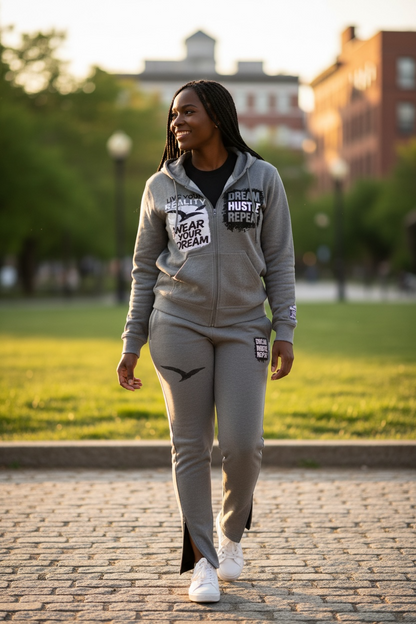 Woman model in my Light grey rhinestone sweatsuit by Je’ Dream Co featuring “Wear Your Dream Live Your Reality” and “Dream Hustle Repeat” graphics, full-zip hoodie and jogger set, premium fabric, sparkling rhinestone details, and luxury streetwear style for everyday motivation in  walking on a brick path with a blurred background