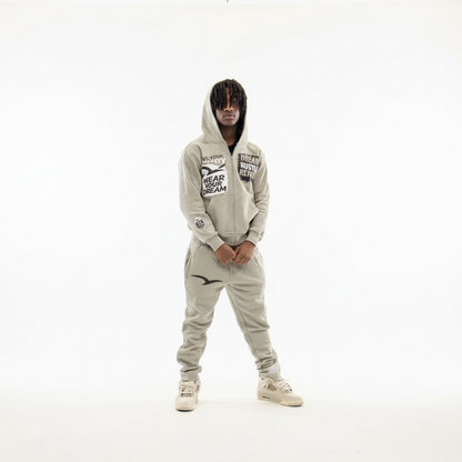 Model wearing    Light grey rhinestone sweatsuit by Je’ Dream Co featuring “Wear Your Dream Live Your Reality” and “Dream Hustle Repeat” graphics, full-zip hoodie and jogger set, premium fabric, sparkling rhinestone details, and luxury streetwear style for everyday motivation on a white background