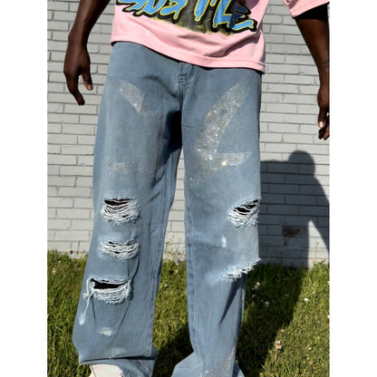 Person wearing our je’ dream co distressed denim blue jeans, bird, feather in rhinestones and a pink shirt with graphic design, standing against a light gray brick wall.